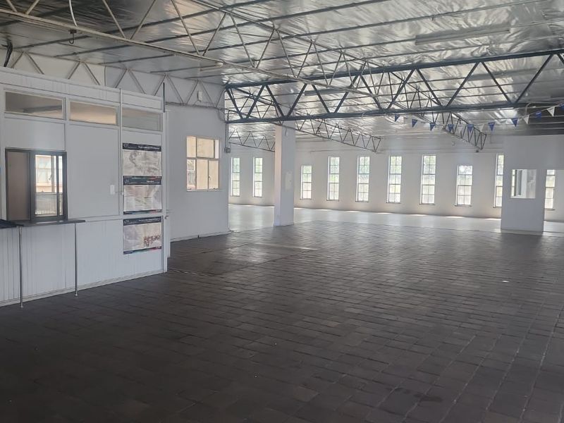 Spacious open-plan industrial unit featuring exposed steel roof trusses, tiled flooring, and a partitioned office booth with natural light from multiple windows.