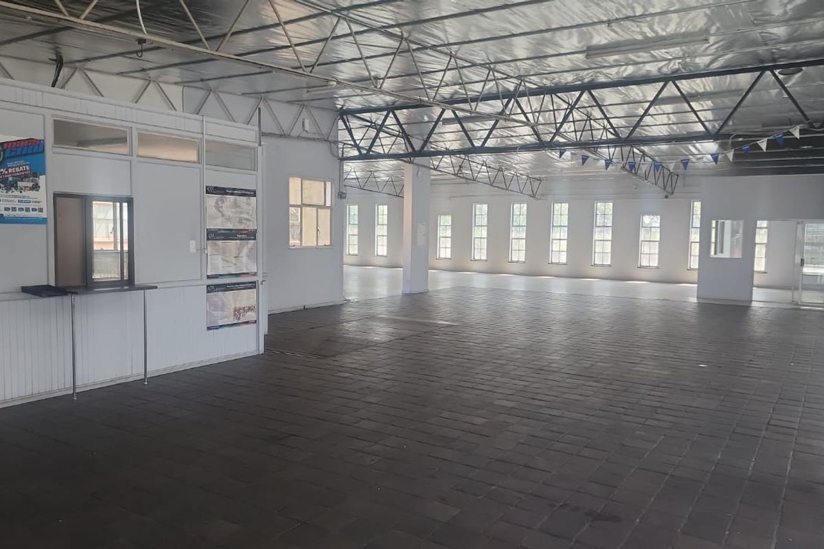 Spacious open-plan industrial unit featuring exposed steel roof trusses, tiled flooring, and a partitioned office booth with natural light from multiple windows.