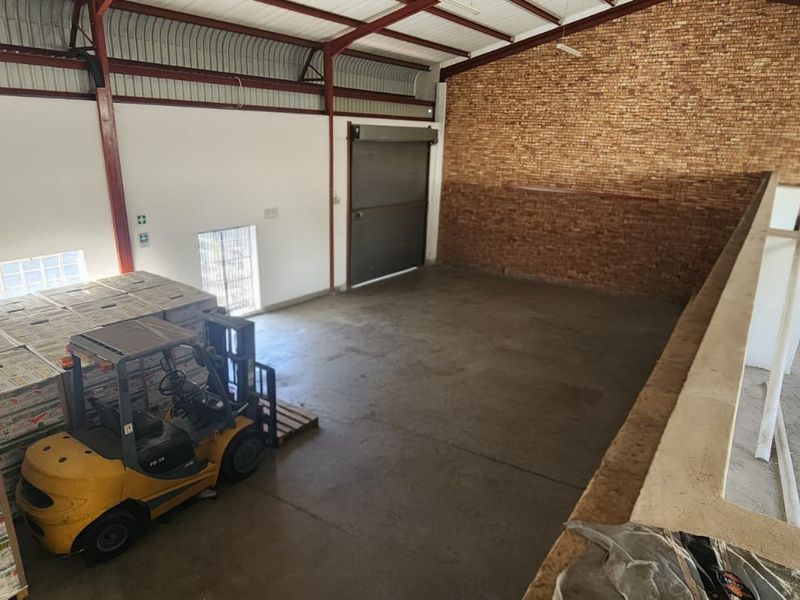 Spacious industrial warehouse area featuring exposed steel roof trusses, concrete flooring, a roller shutter door, and a prominent exposed brick feature wall.