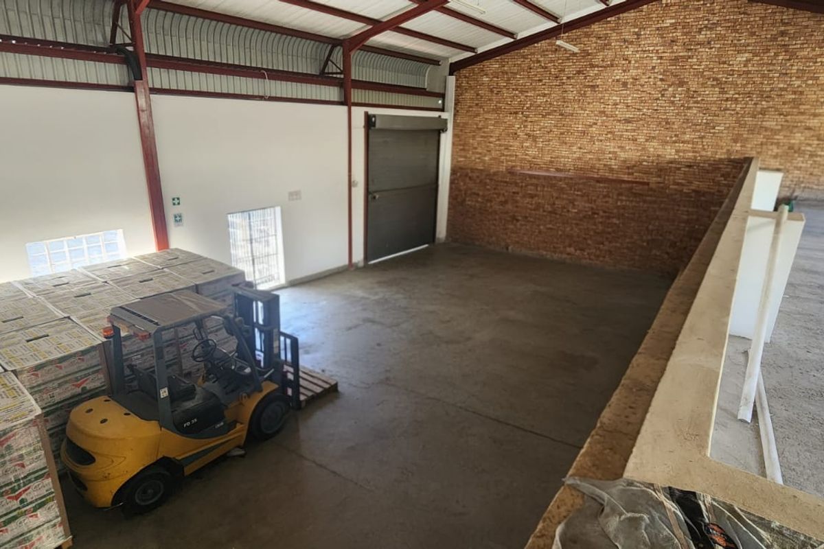 Spacious industrial warehouse area featuring exposed steel roof trusses, concrete flooring, a roller shutter door, and a prominent exposed brick feature wall.