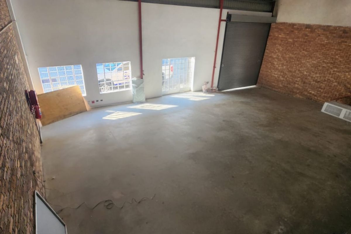 Spacious industrial warehouse unit featuring exposed brick walls, concrete flooring, glass block windows, and a large roller shutter door for easy access.