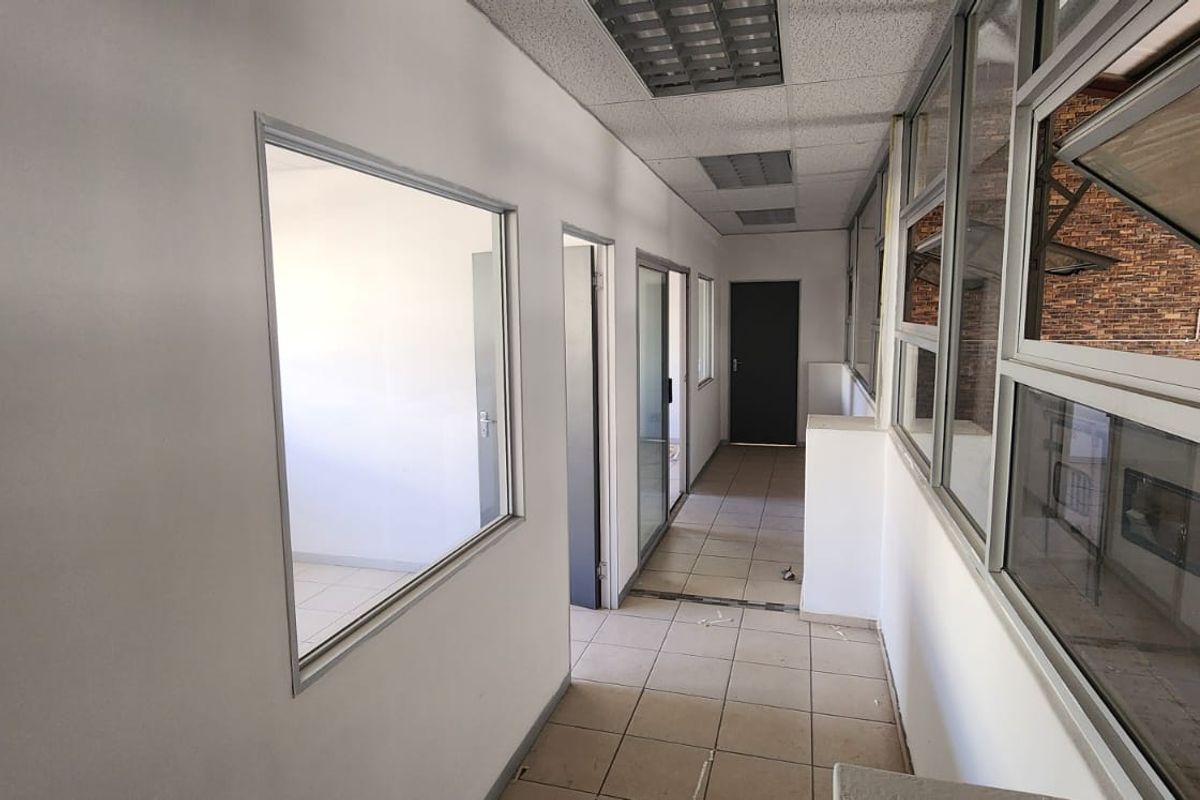 A functional industrial office hallway featuring tiled flooring, suspended ceiling panels, and large glass partitions providing visibility into adjacent workspaces.