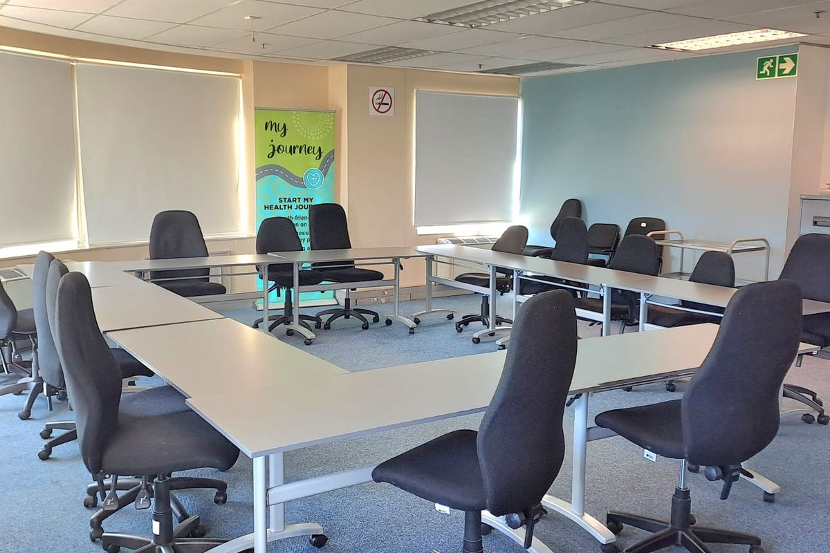 A functional office meeting room featuring a U-shaped desk configuration, suspended ceiling tiles, integrated projector, and roller blinds for light control.