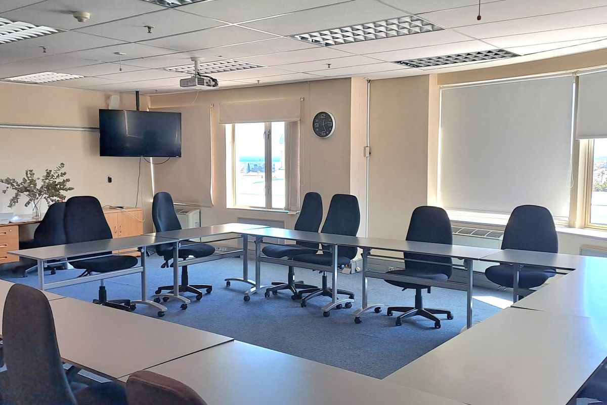 A functional office meeting room featuring a U-shaped desk configuration, suspended acoustic ceiling tiles, recessed lighting, and large windows providing natural light.