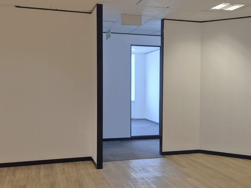 A bright, empty office space featuring neutral walls, laminate flooring, and a suspended ceiling with recessed lighting, leading into an adjacent office area.