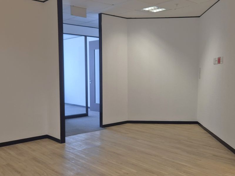 A bright, vacant office space featuring modern laminate flooring, clean white walls, recessed ceiling lighting, and a glass partition door.
