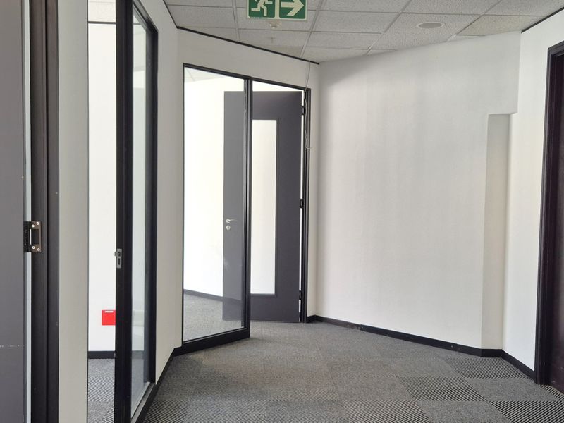 A clean, modern office hallway featuring carpet tiles, suspended ceiling, and glass partition doors leading to individual office spaces.