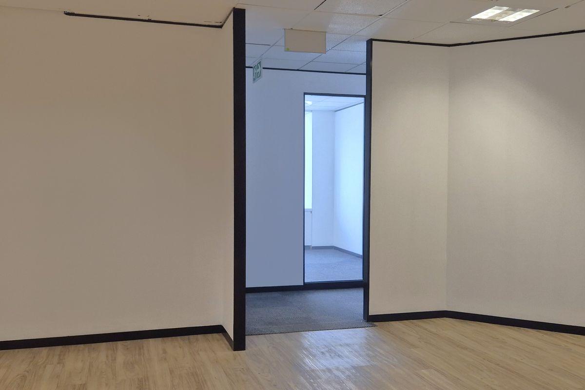 A bright, empty office space featuring neutral walls, laminate flooring, and a suspended ceiling with recessed lighting, leading into an adjacent office area.