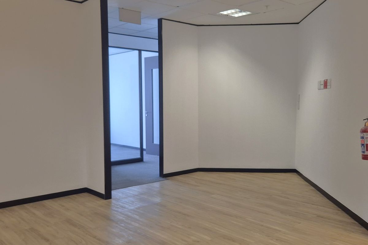 A bright, vacant office space featuring modern laminate flooring, clean white walls, recessed ceiling lighting, and a glass partition door.