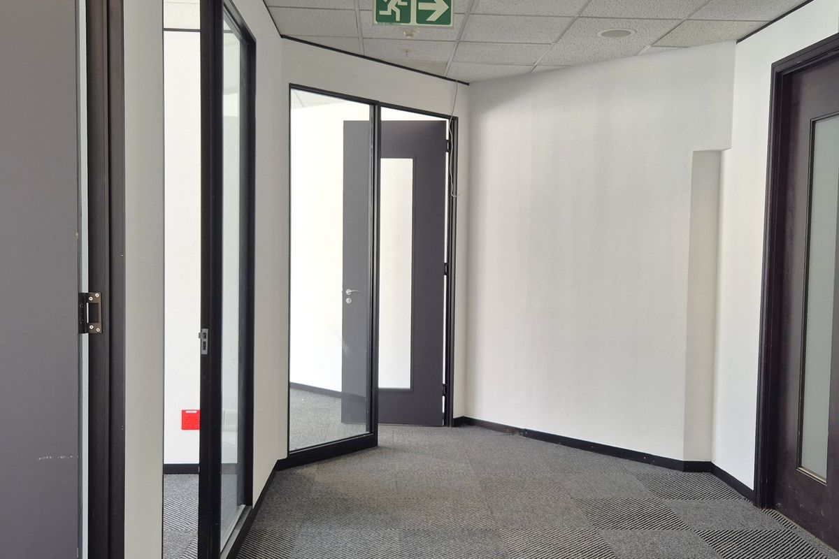 A clean, modern office hallway featuring carpet tiles, suspended ceiling, and glass partition doors leading to individual office spaces.