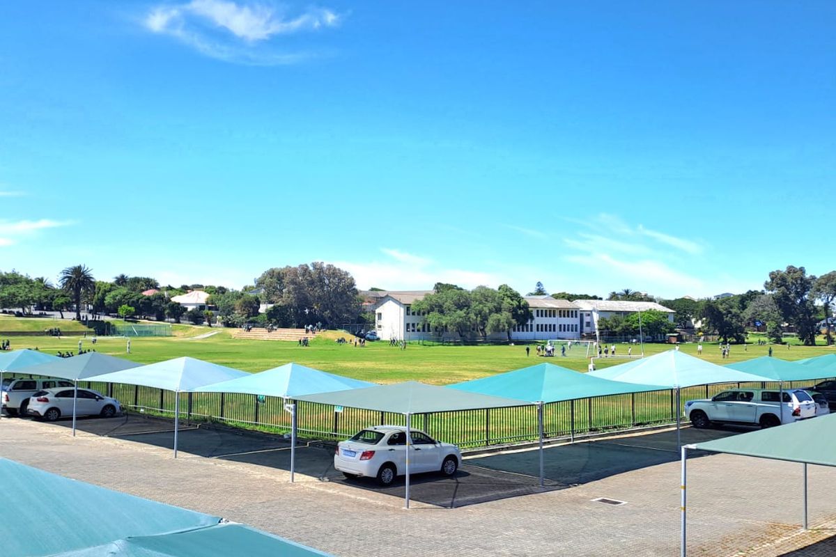 Outdoor parking area featuring multiple shade cloth structures, paved ground, and a view overlooking a large green sports field.