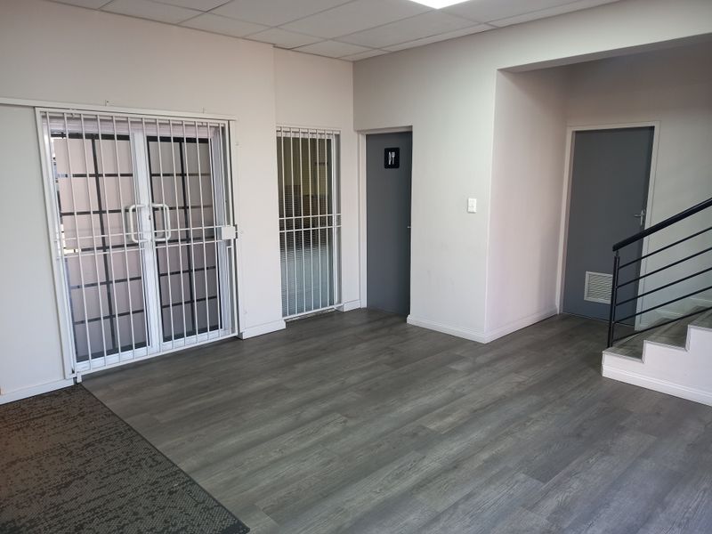 A clean, vacant office space featuring modern grey laminate flooring, secure metal gates, and access to a staircase and restroom facilities.