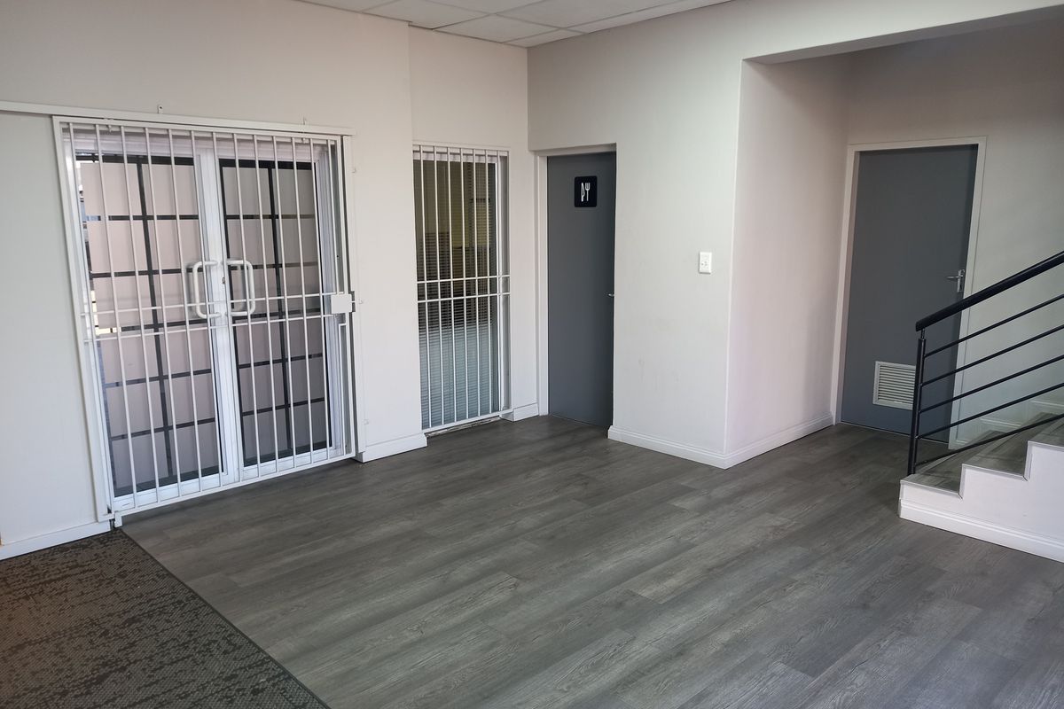 A clean, vacant office space featuring modern grey laminate flooring, secure metal gates, and access to a staircase and restroom facilities.