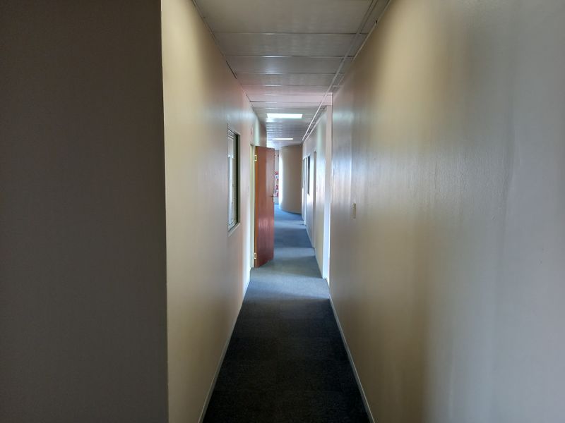 A long office hallway featuring carpeted flooring and a suspended ceiling, providing access to multiple internal office spaces.