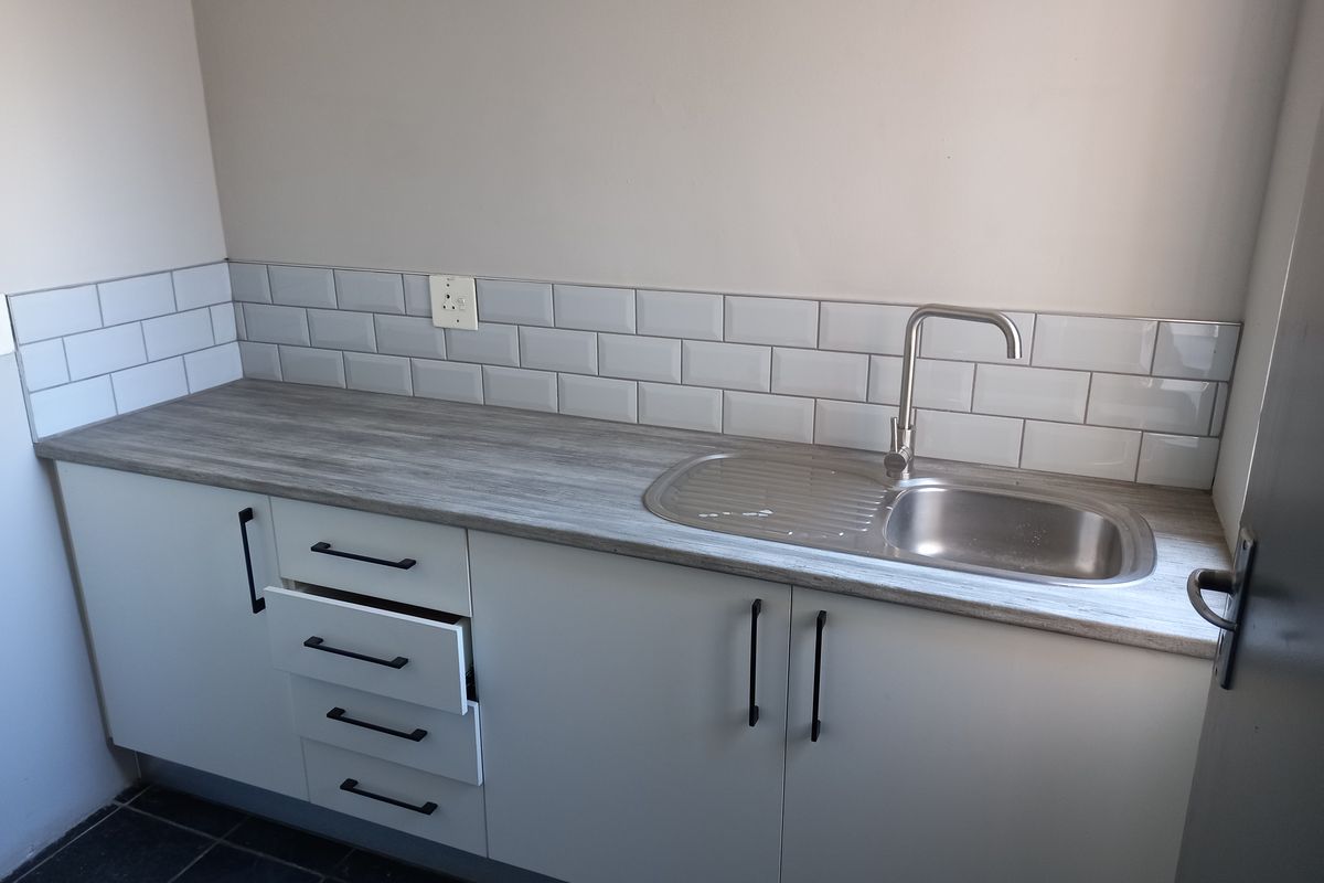 A clean, modern kitchenette area featuring white cabinetry, a wood-look laminate countertop, stainless steel sink, and a stylish subway tile backsplash.