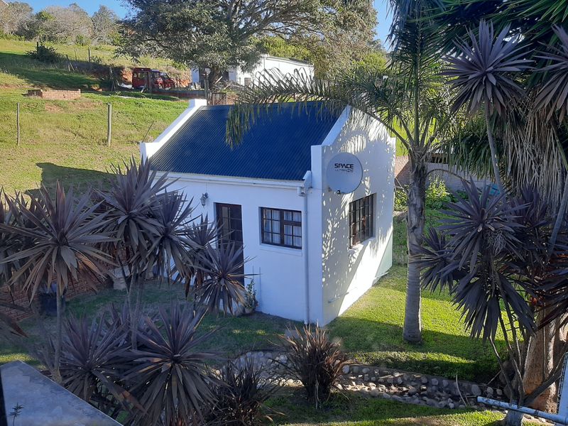A view of a small, white-walled cottage with a dark corrugated metal roof, situated in a landscaped garden setting.