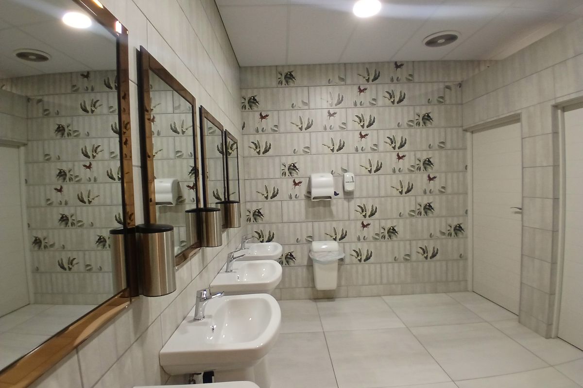 A clean, modern communal bathroom featuring multiple pedestal washbasins, wall-mounted dispensers, and decorative tiled walls with neutral floor tiling.
