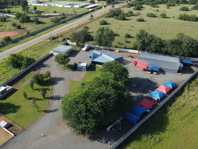 Aerial view of a commercial property featuring a large warehouse, multiple shade cloth parking bays, gravel yard, and landscaped garden areas.