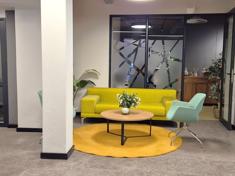 A modern office breakout area featuring a glass partition wall, neutral flooring, and professional lighting fixtures.