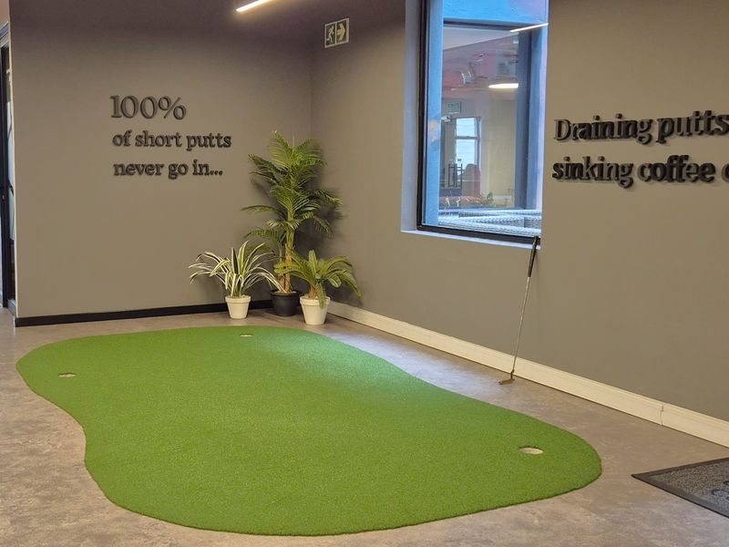 Modern office breakout area featuring an indoor putting green, glass-partitioned corridors, and contemporary grey wall finishes with exposed ceiling infrastructure.