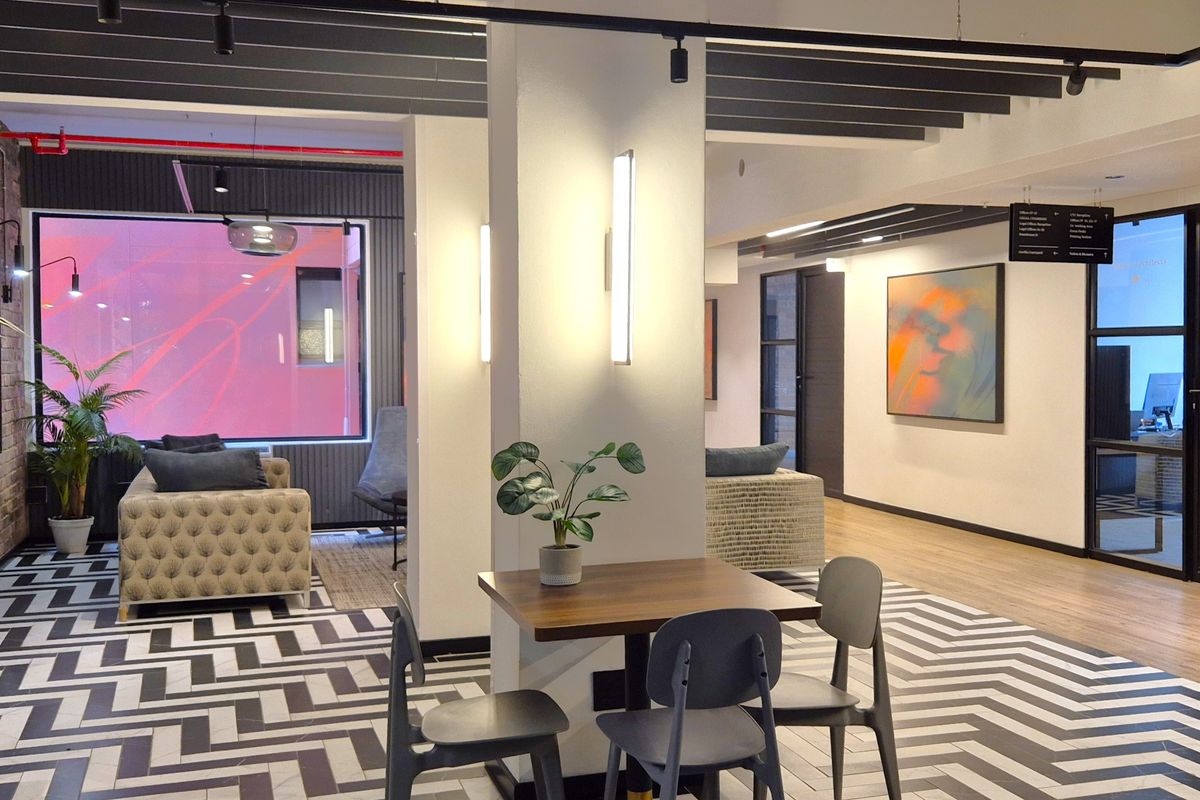 Modern open-plan office breakout area featuring a mix of herringbone tile and laminate flooring, exposed ceiling beams, and contemporary track lighting.