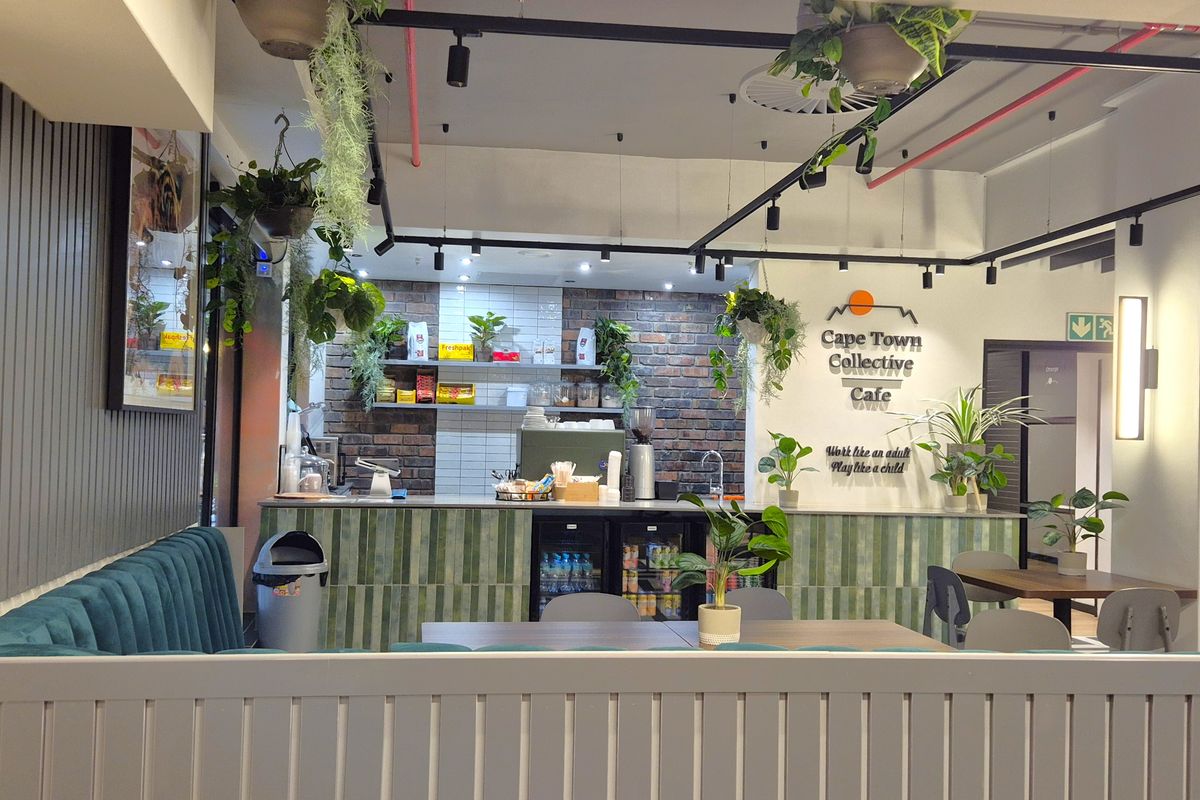 Modern open-plan office cafe area featuring a brick accent wall, service counter with integrated refrigeration, and industrial-style exposed ceiling infrastructure.