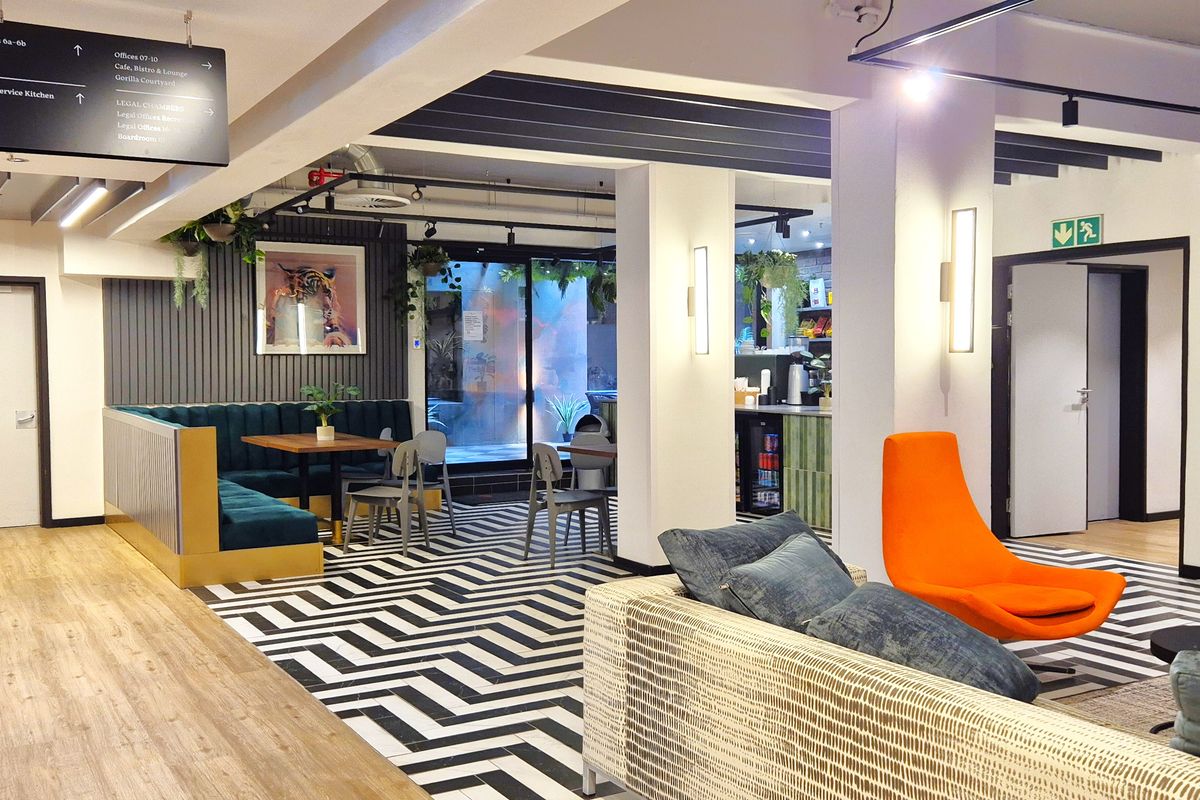 Modern open-plan office breakout area featuring a mix of wood-look flooring, patterned tiling, exposed brick accents, and integrated wayfinding signage.