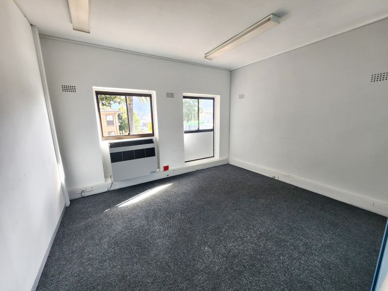 An empty office space featuring grey carpet flooring, dual sliding windows, and a built-in air conditioning unit.