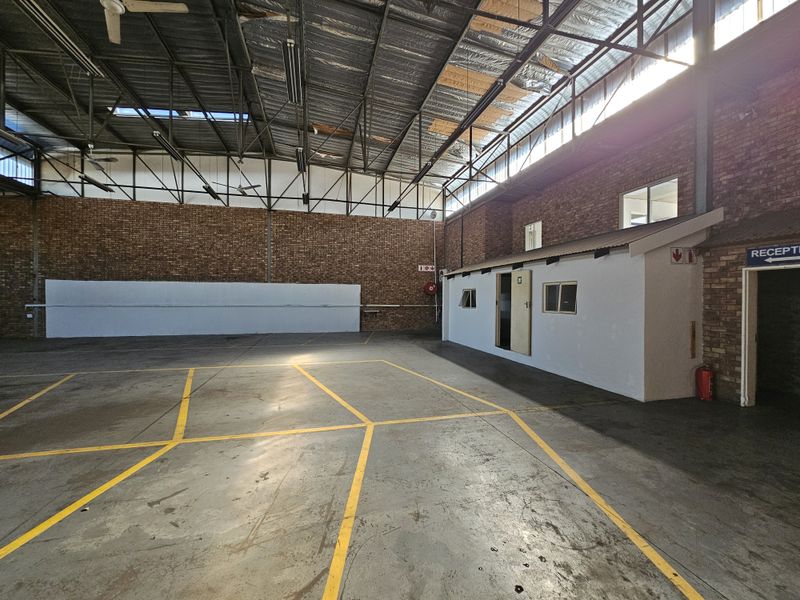 Spacious industrial warehouse area featuring exposed brick walls, high steel roof trusses, concrete flooring with marked bays, and an integrated office structure.