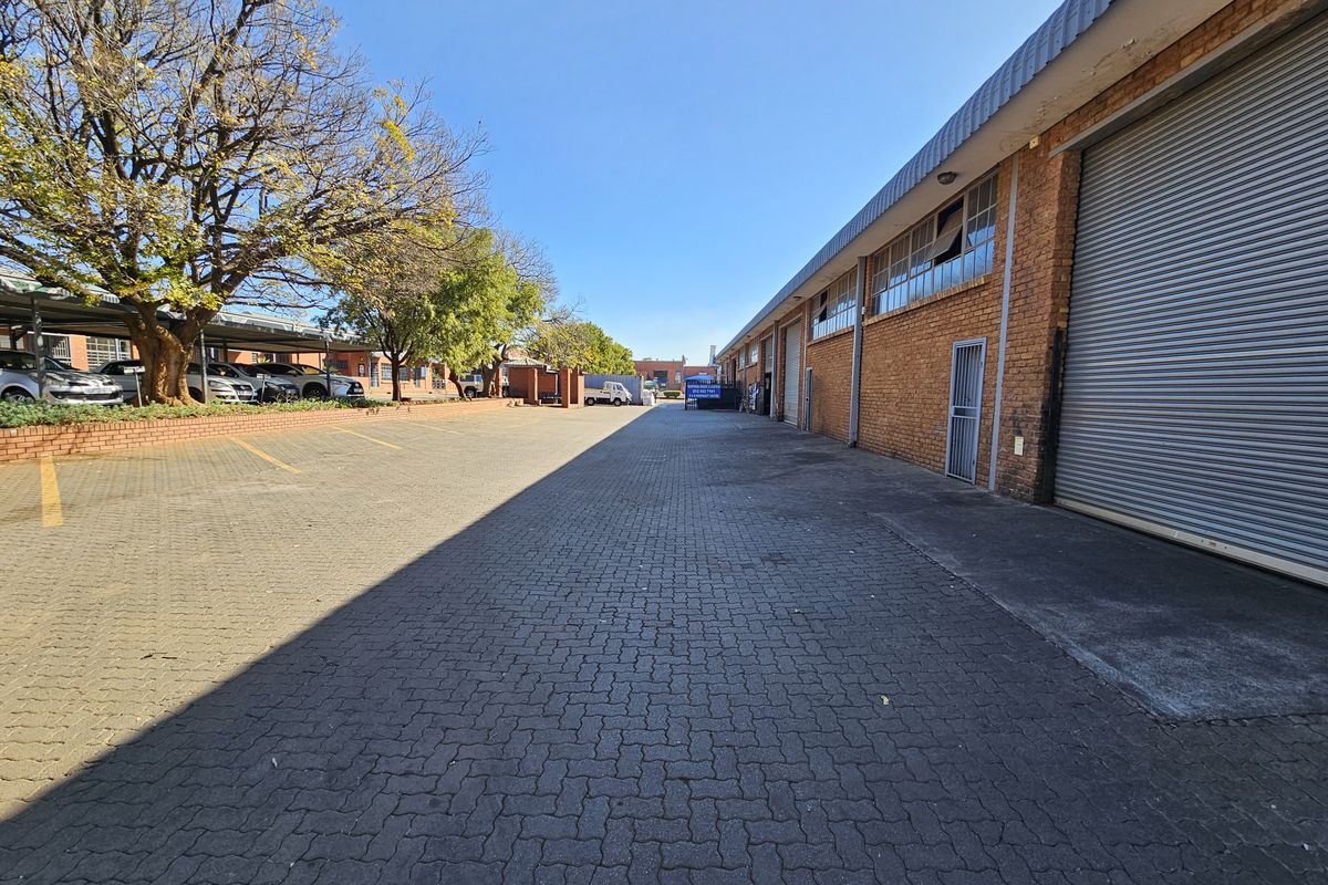 Industrial exterior view featuring a paved driveway, brick facade, and multiple roller shutter doors with adjacent covered parking bays.
