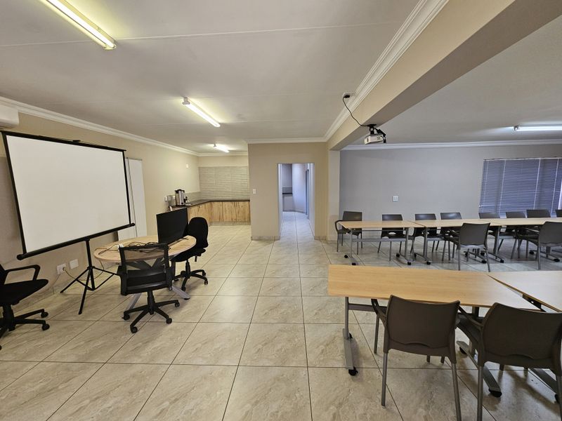 Spacious office or conference room featuring tiled flooring, integrated kitchenette area, ceiling-mounted projector, and ample natural light through venetian blinds.