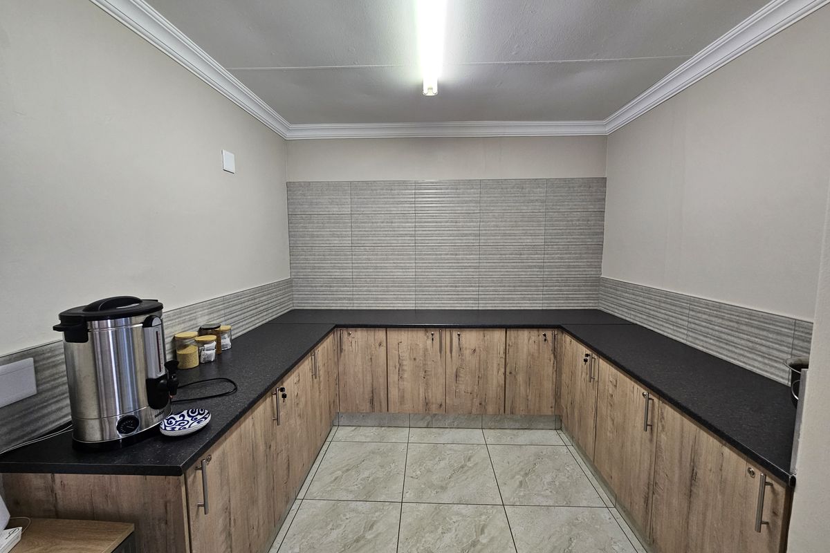 A clean and functional kitchenette area featuring modern wood-look cabinetry, dark countertops, and neutral tiled flooring, suitable for office staff use.