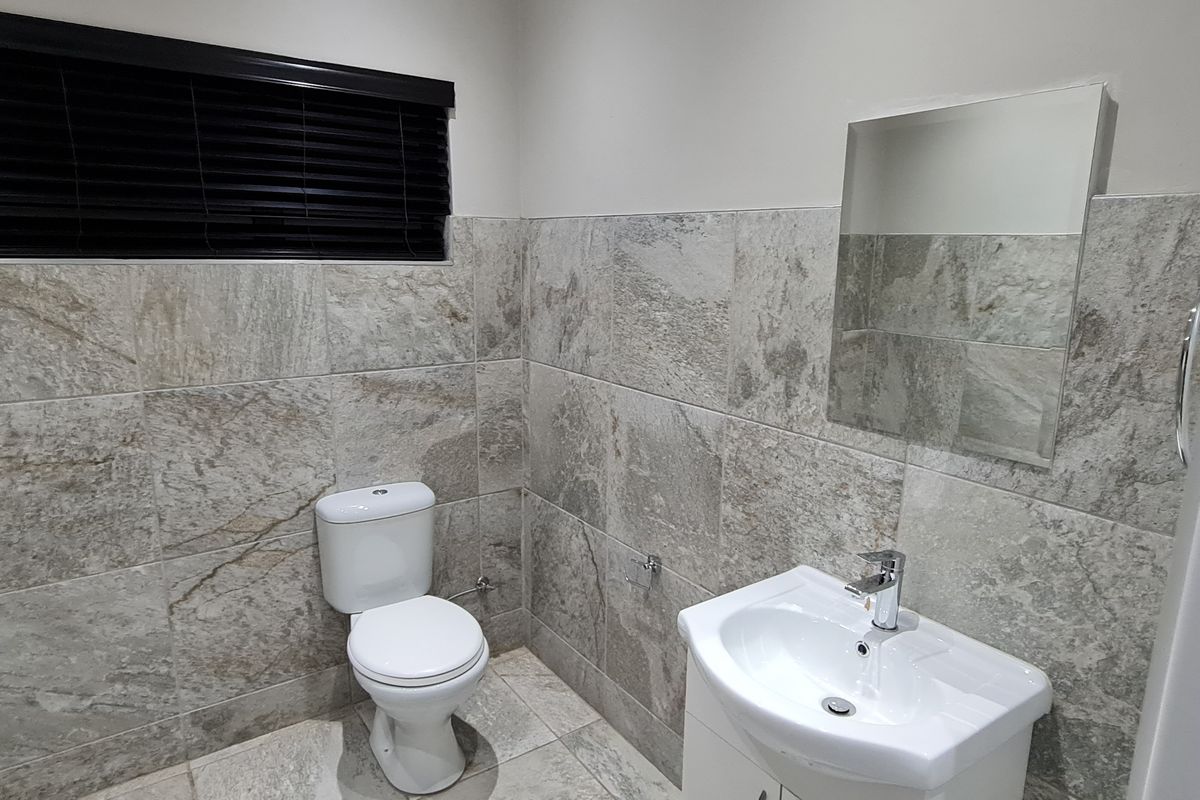 A modern office bathroom featuring floor-to-ceiling stone-look wall tiles, a white pedestal basin, a wall-mounted mirror, and a sleek toilet.
