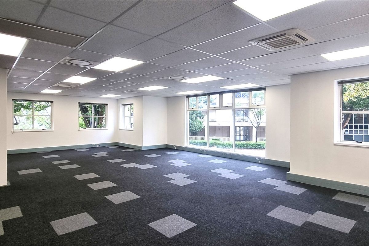 A spacious, open-plan office area featuring modern carpet tiles, recessed lighting, a suspended ceiling, and large windows providing ample natural light.