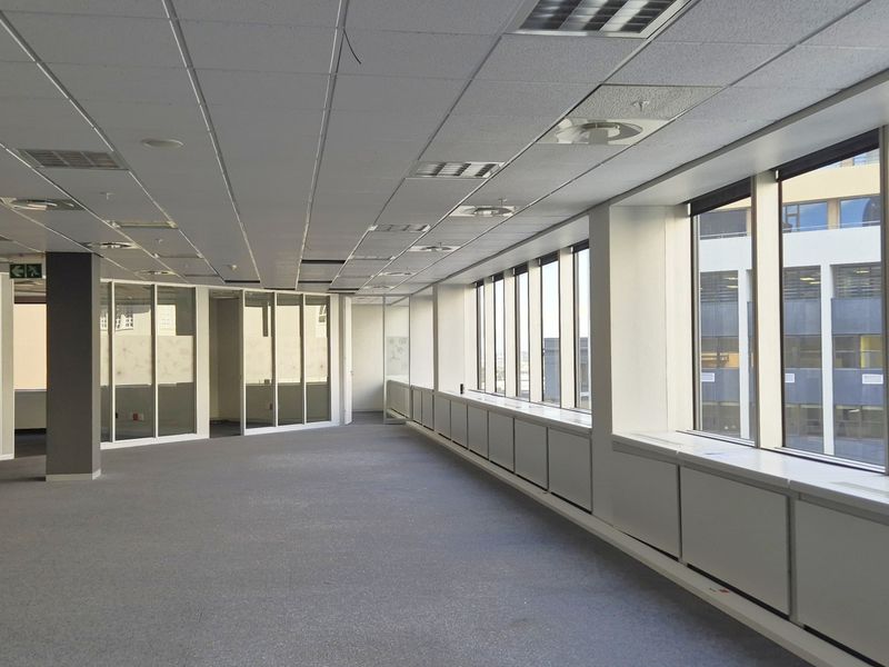 A spacious, vacant open-plan office area featuring large windows, glass partitions, carpeted flooring, and a suspended ceiling with integrated lighting and air conditioning.