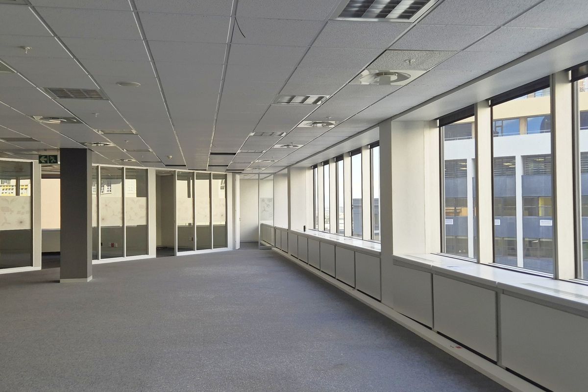 A spacious, vacant open-plan office area featuring large windows, glass partitions, carpeted flooring, and a suspended ceiling with integrated lighting and air conditioning.
