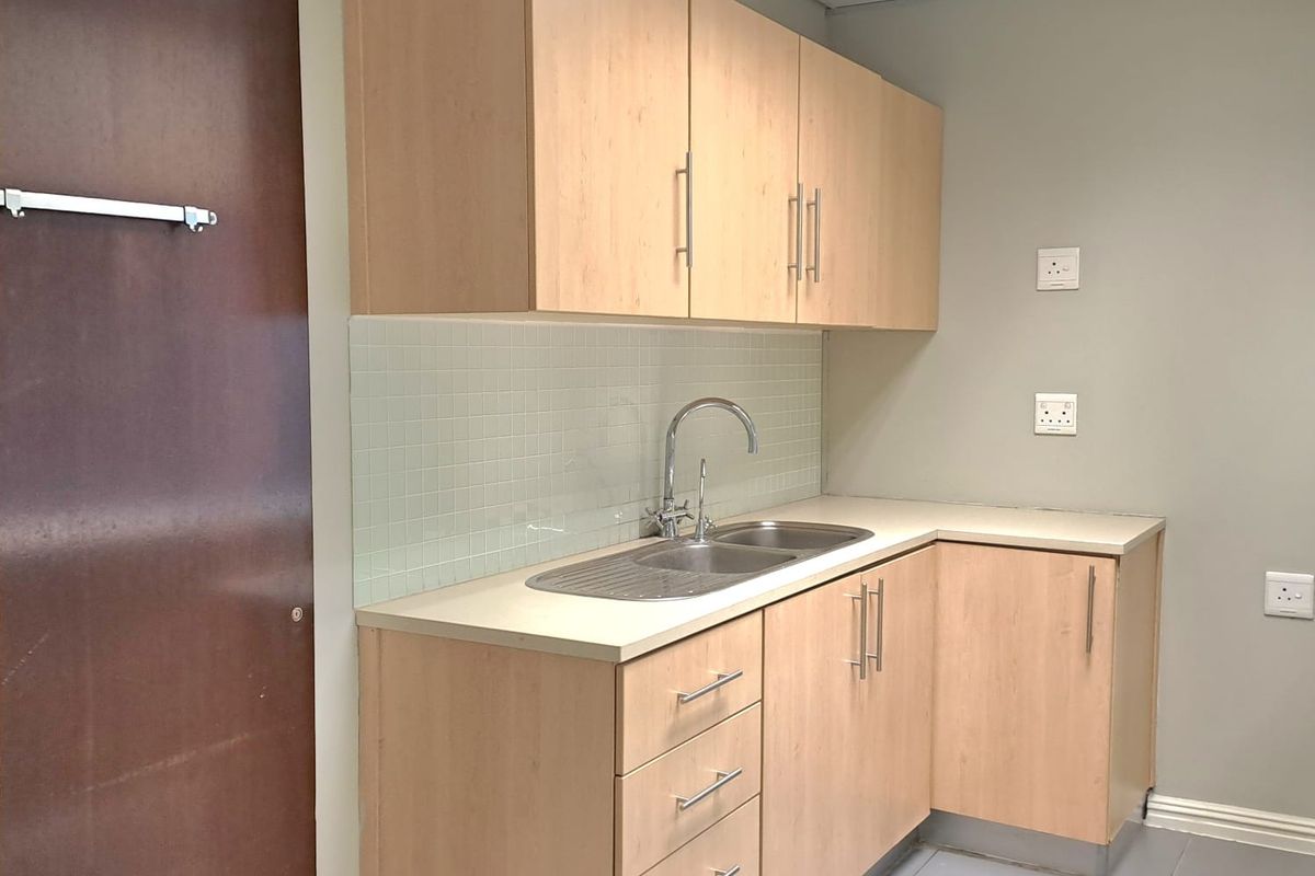 A functional office kitchenette featuring light wood cabinetry, a stainless steel sink, tiled backsplash, and grey floor tiles.