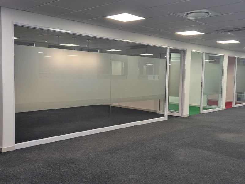 Modern open-plan office space featuring glass-partitioned private offices, recessed ceiling lighting, air conditioning units, and carpeted flooring throughout.