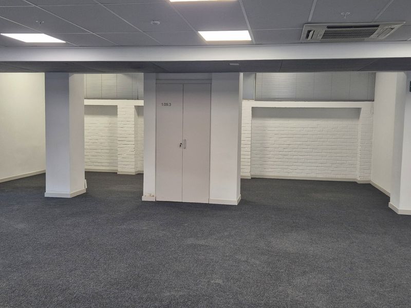 An open-plan office space featuring a suspended ceiling with recessed lighting, central support columns, and dark carpeting.