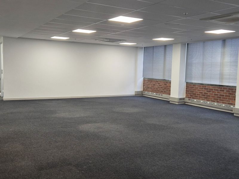 Spacious open-plan office area featuring suspended ceiling tiles, recessed LED lighting, integrated air conditioning units, and large windows with brickwork detailing.