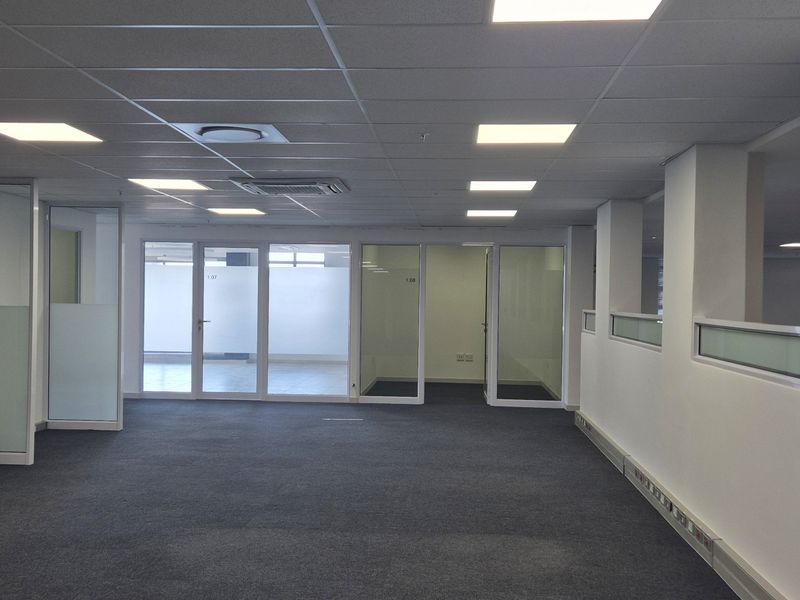 A spacious, open-plan office area featuring a suspended ceiling with recessed lighting, glass-partitioned offices, and carpeted flooring.