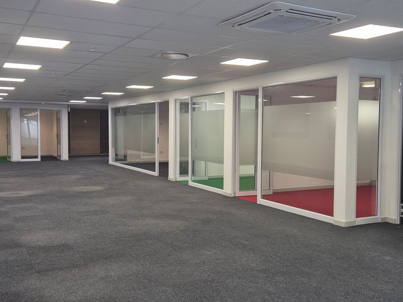 A spacious, open-plan office area featuring modern glass-partitioned meeting rooms, recessed ceiling lighting, and carpeted flooring throughout.