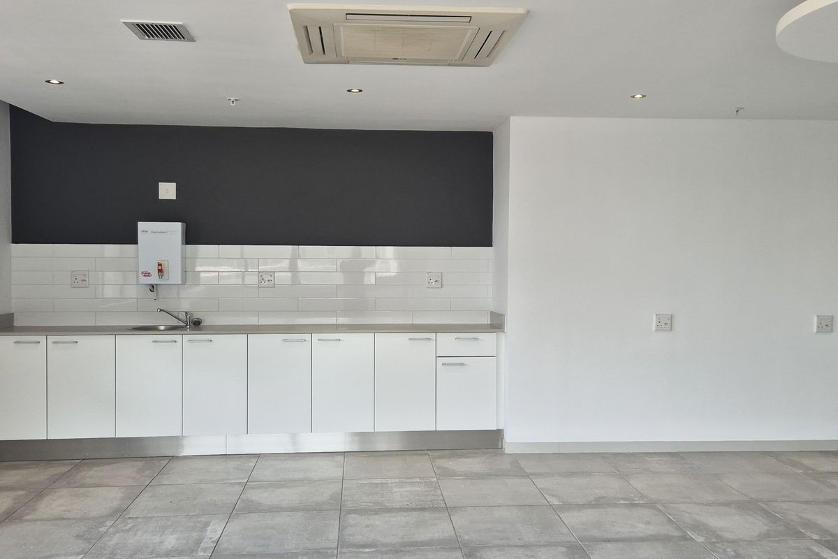 A clean, modern office kitchenette featuring white cabinetry, subway tile backsplash, stainless steel sink, and integrated ceiling air conditioning unit.