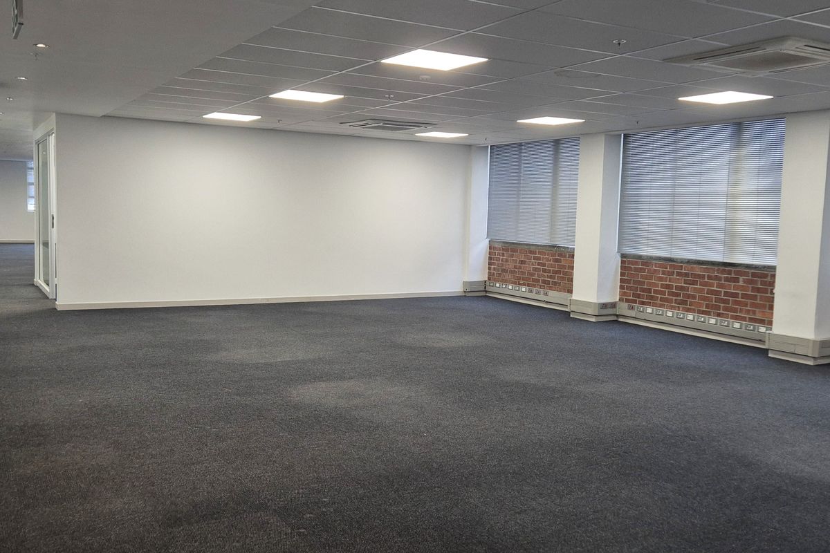 Spacious open-plan office area featuring suspended ceiling tiles, recessed LED lighting, integrated air conditioning units, and large windows with brickwork detailing.