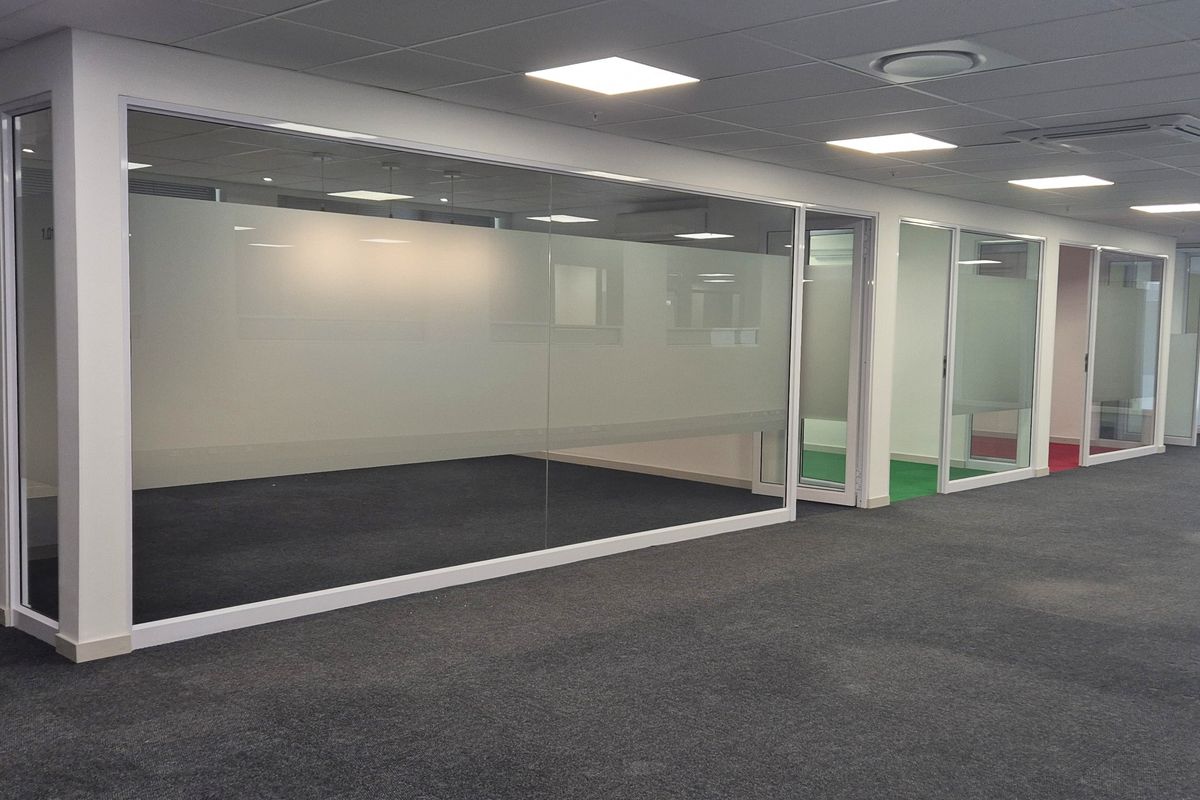 Modern open-plan office space featuring glass-partitioned private offices, recessed ceiling lighting, air conditioning units, and carpeted flooring throughout.