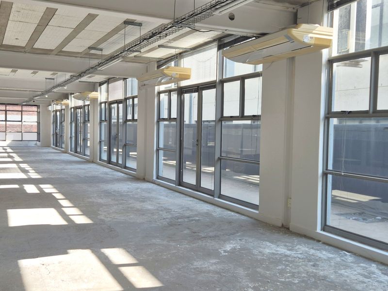 Spacious open-plan office area featuring exposed concrete ceilings, large windows providing natural light, and wall-mounted air conditioning units.