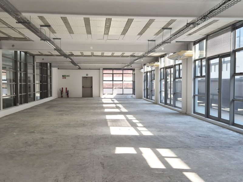 Spacious open-plan office area featuring industrial-style exposed ceilings, concrete flooring, and extensive natural light through large perimeter windows.