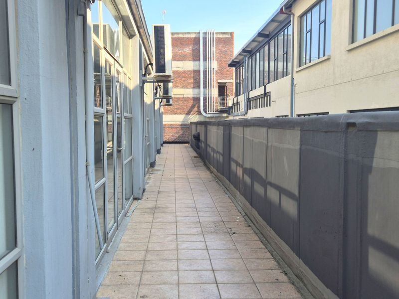An exterior balcony walkway featuring tiled flooring, aluminum-framed windows, and an external air conditioning unit, providing access between office building sections.