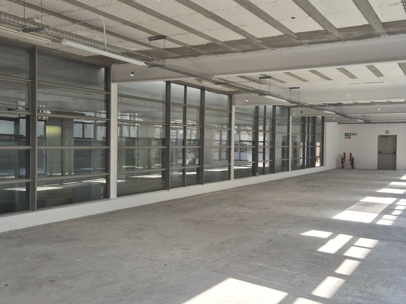 Spacious open-plan office area featuring industrial-style exposed concrete ceilings, large glass partitions, and polished concrete flooring, suitable for a modern workspace layout.
