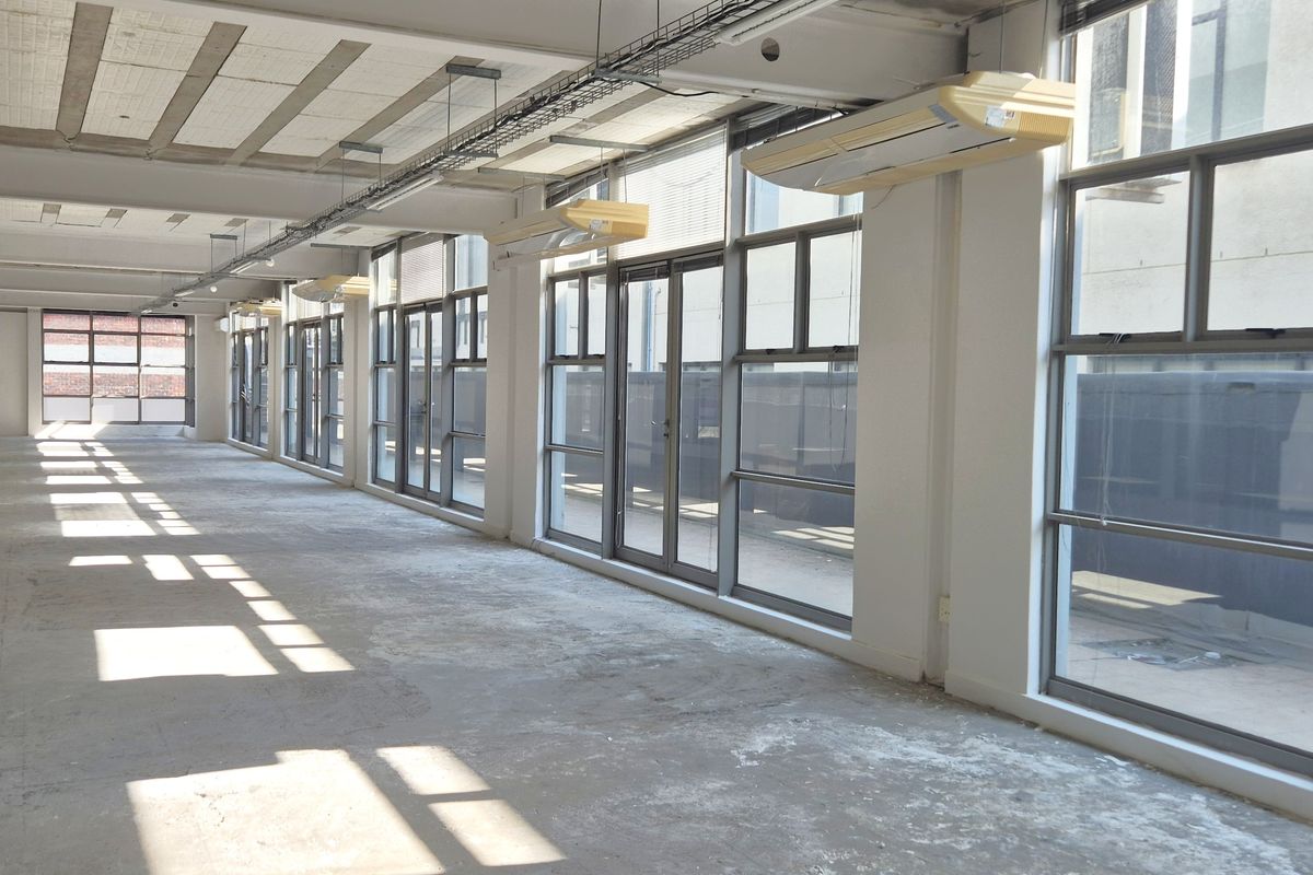Spacious open-plan office area featuring exposed concrete ceilings, large windows providing natural light, and wall-mounted air conditioning units.